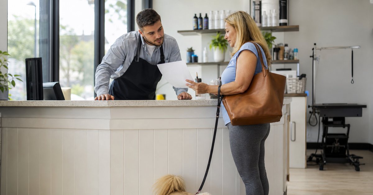 A groomer in an apron at a bright salon front counter looking at a paper document held by a woman with a small fluffy dog on a leash, morning light through the window